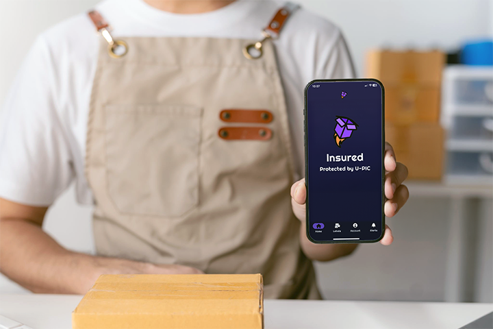 Image: An eCommerce seller holds up a smartphone displaying the I’d Ship That app, showing the U-PIC insured badge.