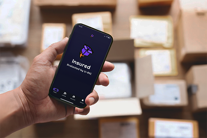 Image: An eCommerce seller holds up a smartphone above shipping boxes displaying the I’d Ship That app, showing the U-PIC insured badge.