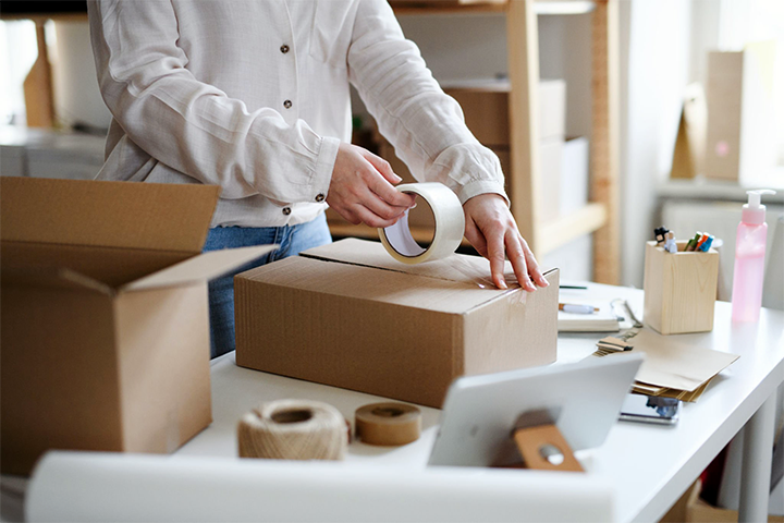 Image: Person sealing a cardboard box with packing tape on a desk, representing proper packaging practices for shipping.