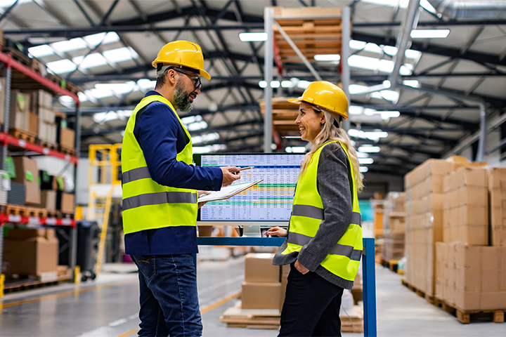 Image: Two warehouse workers strategizing logistics in front of a digital screen.