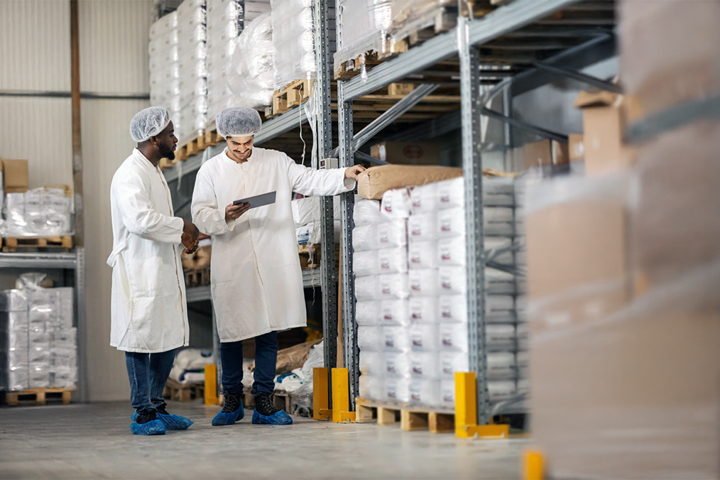 Image: Two warehouse workers in sterile clothing looking over packages for shipping.