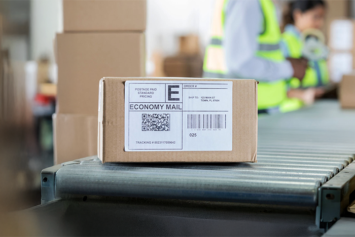 Image: A box with a shipping label sitting on a conveyor belt in a package warehouse.