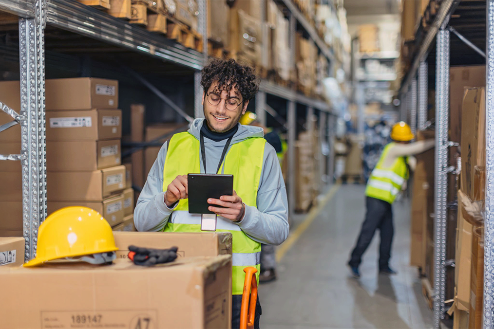 Image: A warehouse worker reviewing outbound orders on a tablet next to a stacked pallet of boxes.